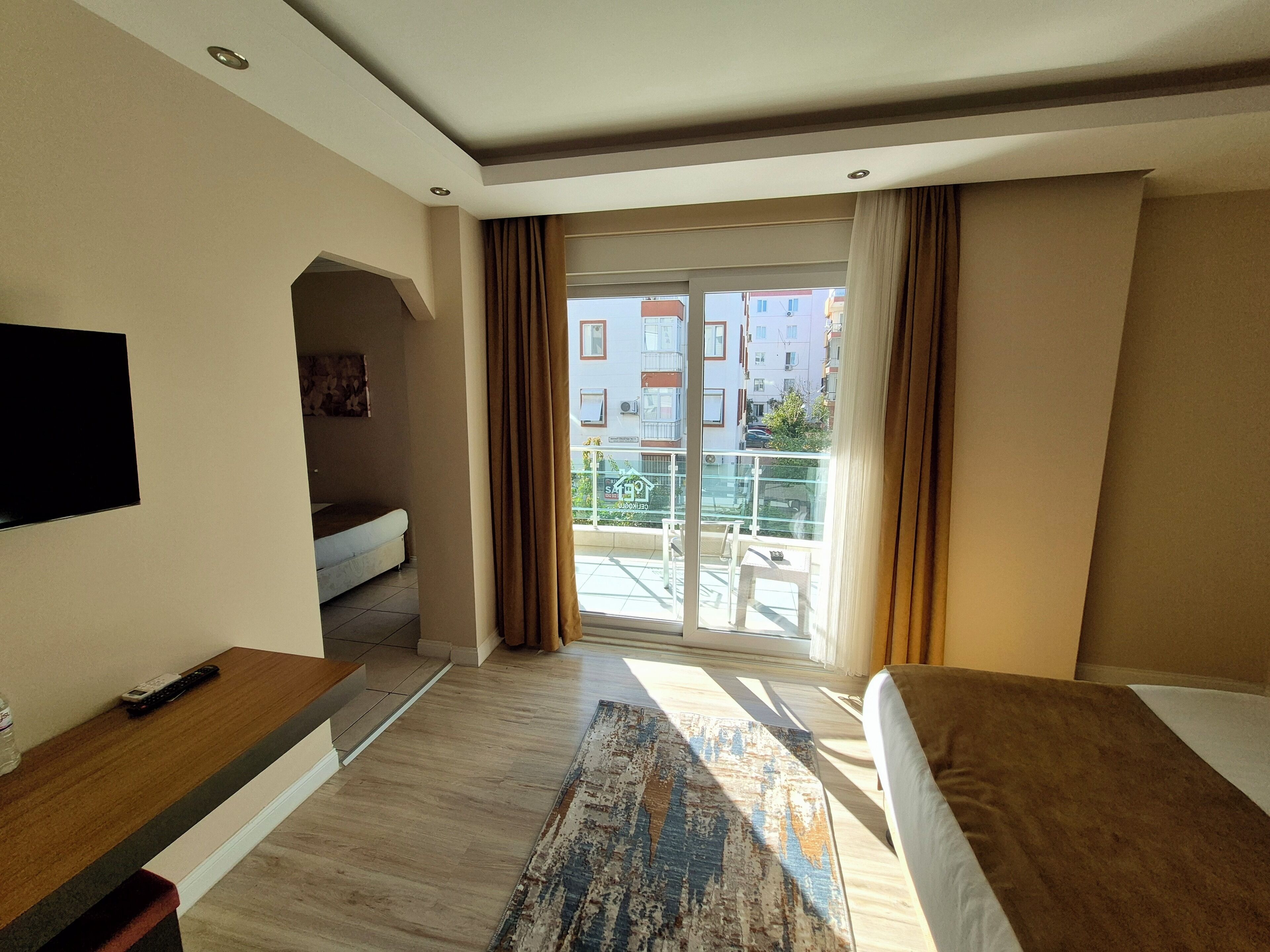 Habitació d'hotel amb un llit, una televisió, un balcó amb vistes a edificis i una taula petita amb un comandament a distància.