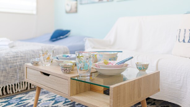 A neatly arranged dining area with a glass-top table, various bowls and cups, and a bed in the background.