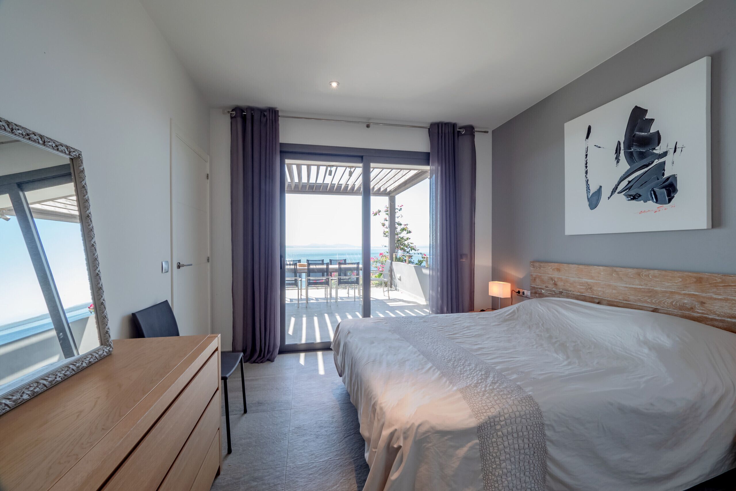 A hotel room with a bed, a dresser, a mirror, a window with curtains, and a view of the sea.