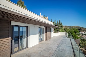 Un edificio moderno con grandes puertas correderas de vidrio, un balcón con suelo de piedra y vistas a un paisaje costero.
