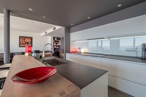 A modern kitchen with white cabinetry, a wooden countertop, and a red bowl. There is a dining area with a long table and a sofa. A bookshelf and a lamp are visible in the background. - VILLA VELAZQUEZ Roses (Rosas / Roses)