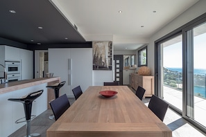 Eine moderne Küche mit einem Holztisch, schwarzen Barhocker und einem großen Fenster mit Blick auf das Meer.