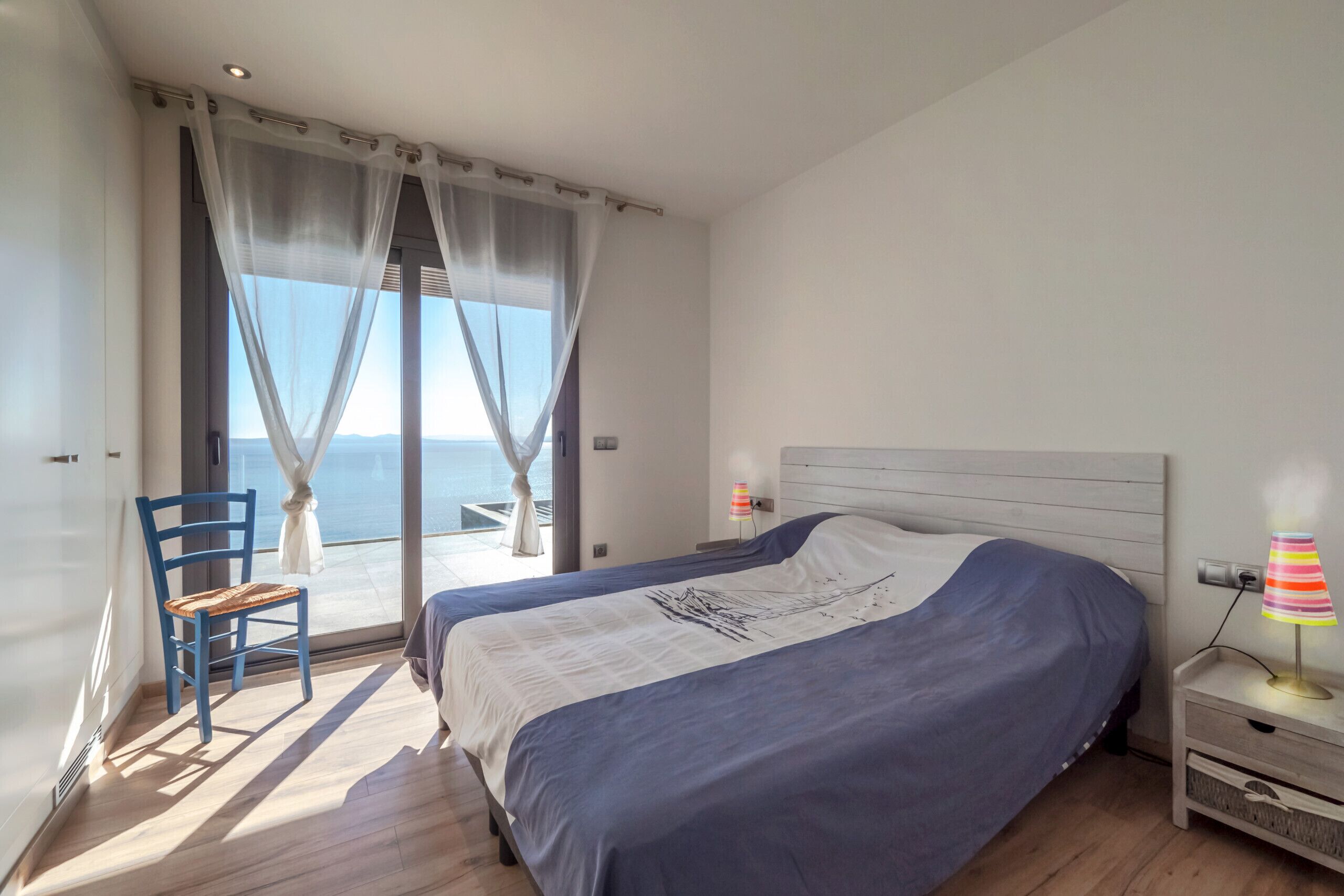 Slaapkamer met een groot bed, een stoel, een nachtkastje en uitzicht op de zee door dunne gordijnen.