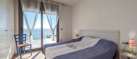 Slaapkamer met een groot bed, een stoel, een nachtkastje en uitzicht op de zee door dunne gordijnen.
