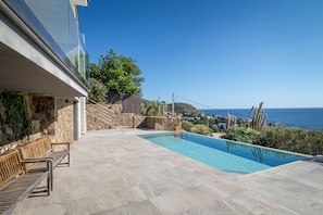 Zona de piscina exterior moderna con vista despejada al mar, paredes de piedra y bancos de madera.