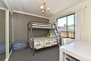 A bunk bed with a patterned bedspread, a desk, a chair, a bean bag, and a window with a view of a house.