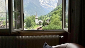A room with a large window offering a view of a mountainous landscape, a chair with a pillow, and a radiator. - THE Best Valley View •  Swiss Charm • Free Parking (Lauterbrunnen)
