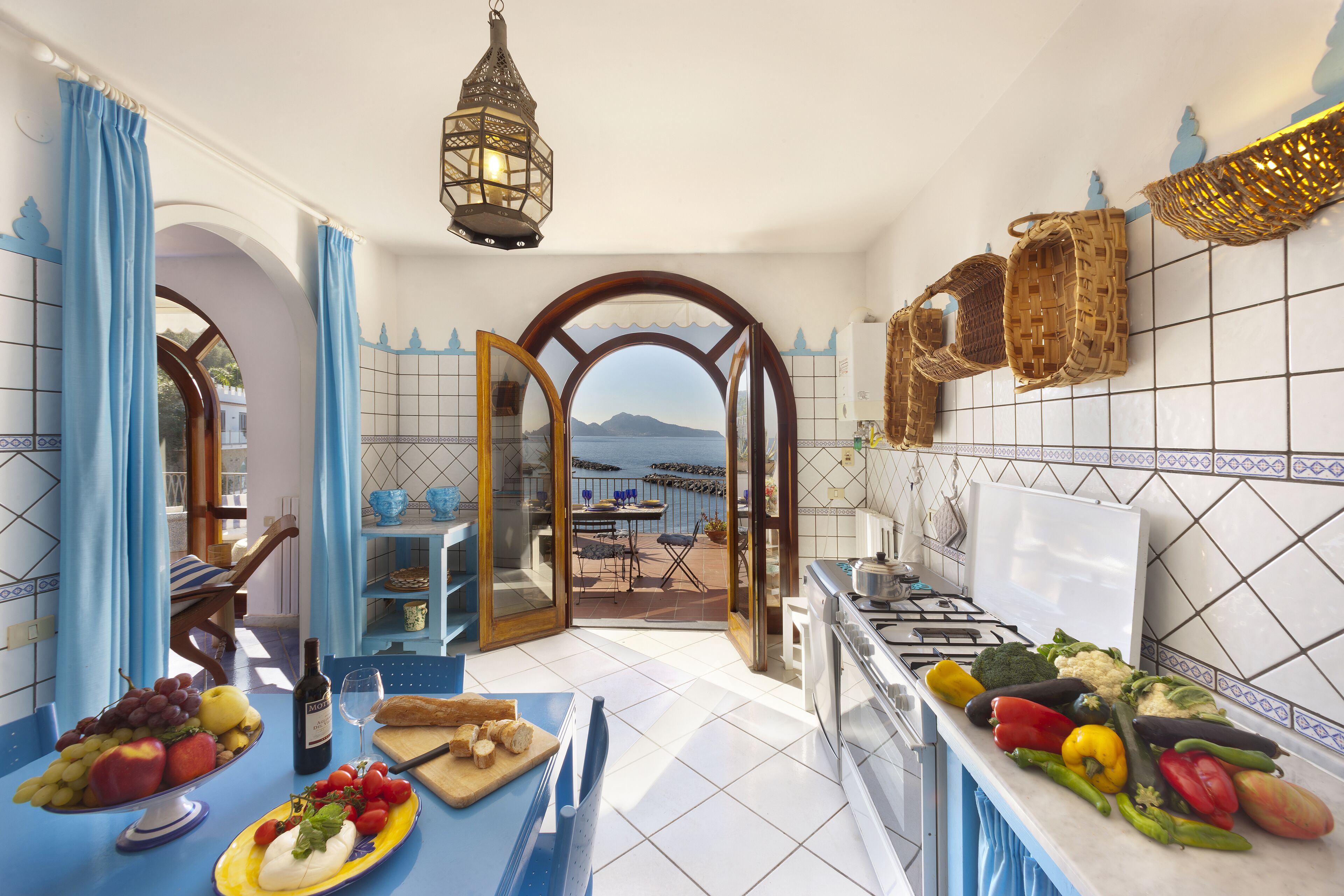 A kitchen with a view of the sea, a dining area with blue curtains, and a variety of fruits and vegetables on the counter.