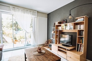 Uma sala de estar moderna com uma televisão grande, estantes de madeira, uma mesa de jantar e vista para um jardim através de janelas amplas.