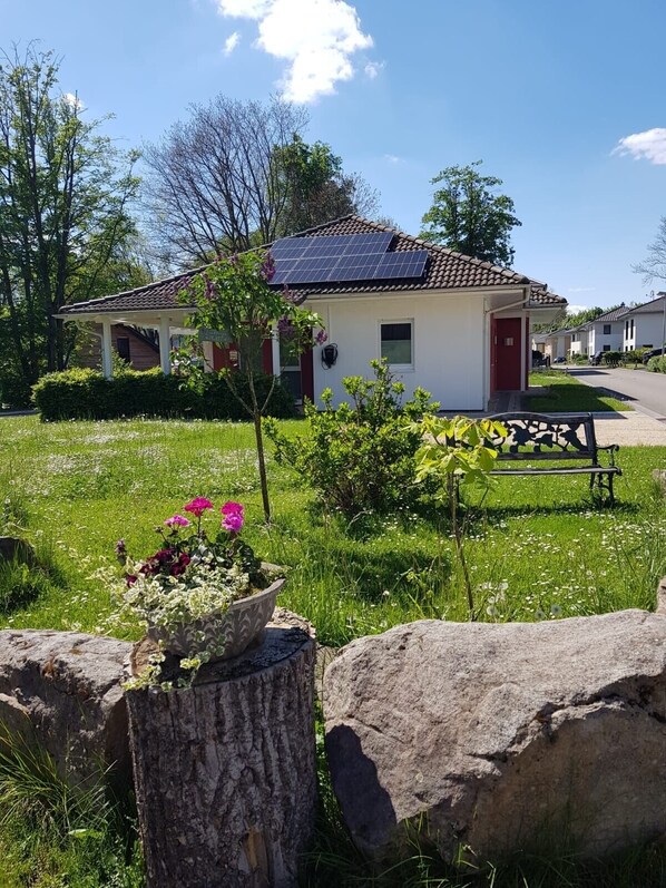 Een gelijkvloers huis met pannendak, zonnepanelen en een kleine tuin met bloemen en een bankje.