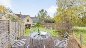 A wooden deck with metal chairs and a table set for dining, surrounded by a stone wall and a garden.