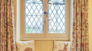 A window with a diamond-patterned grille, a bench with two cushions featuring hedgehogs, and floral curtains.