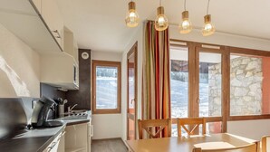 A compact kitchen with a stove, sink, and cabinets, a wooden dining table with chairs, and large windows with a view of a snowy landscape.
