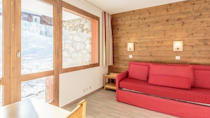 A room with a wooden wall, a red sofa, a wooden table, and a large window with a stone wall view.