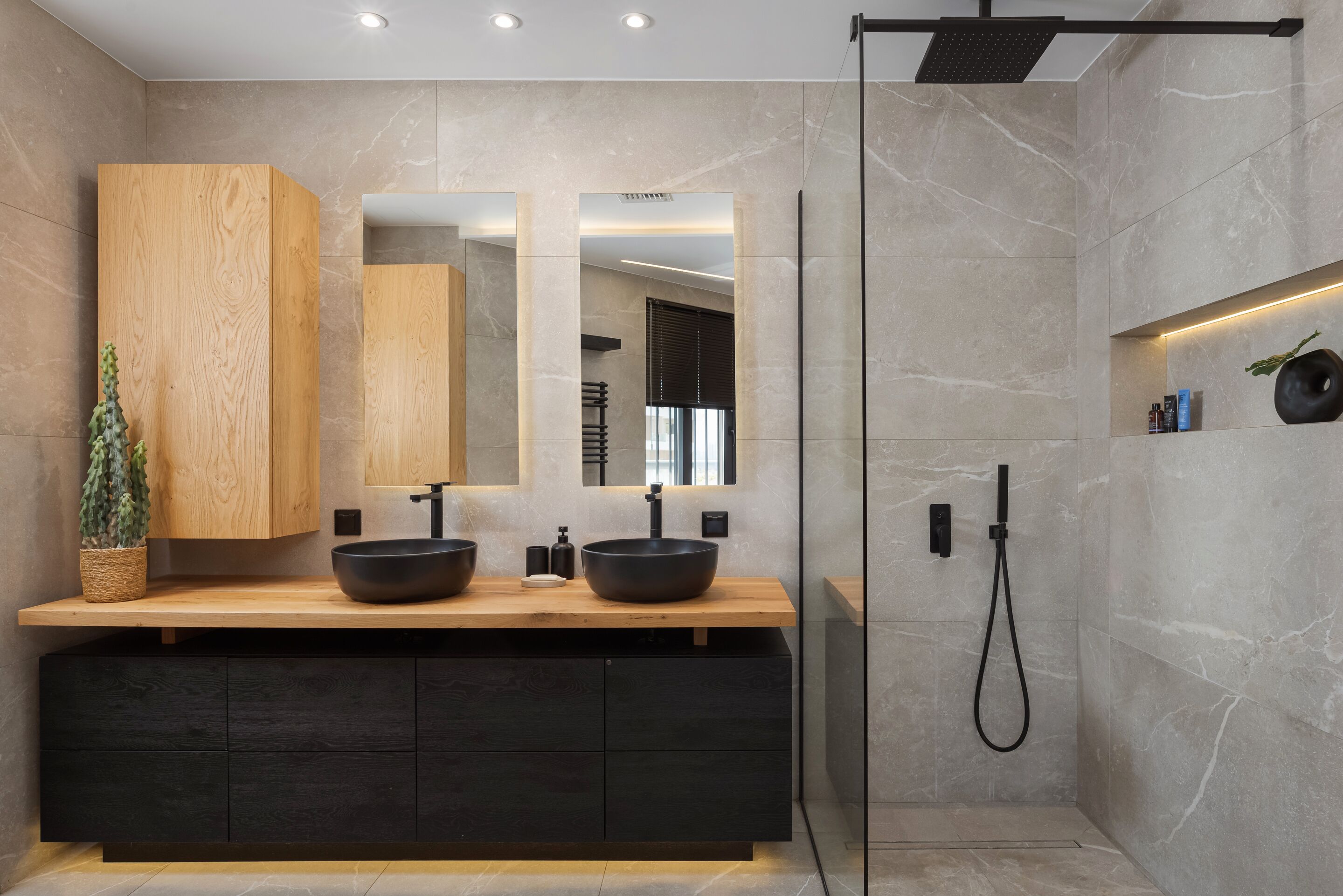 A modern bathroom with a wooden vanity, two black vessel sinks, a large mirror, and a shower area.