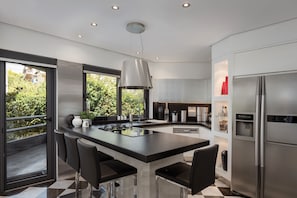 A modern kitchen with a central island, stainless steel appliances, and a view of greenery outside. - Athenian Riviera Luxurious Villa & Residence (Alimos)