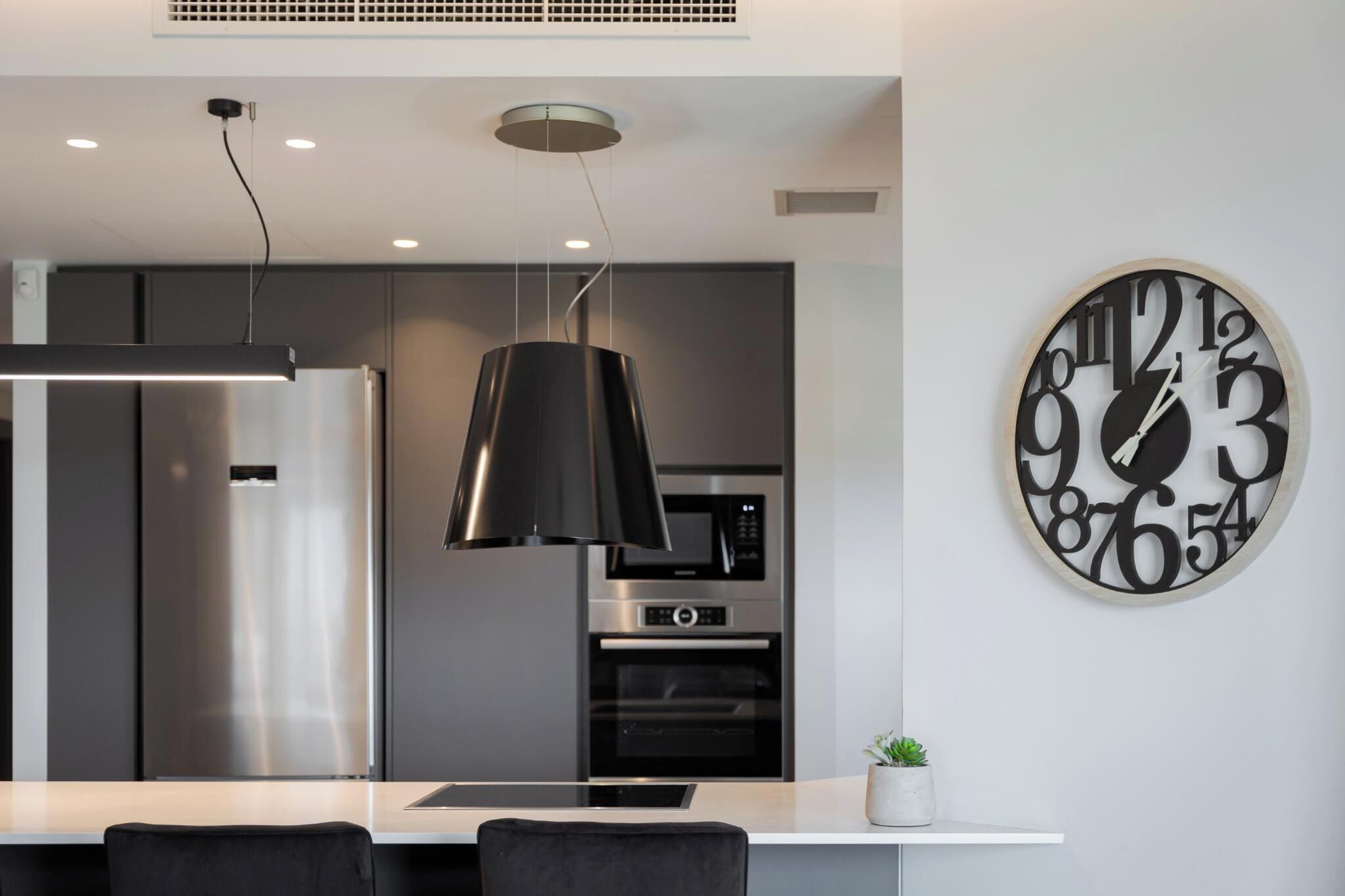 Une cuisine moderne équipée d’un réfrigérateur en acier inoxydable, d’un four à micro-ondes et d’un four traditionnel. Un grand réveil mural et un luminaire suspendu ornent le mur au-dessus du plan de travail de la cuisine.