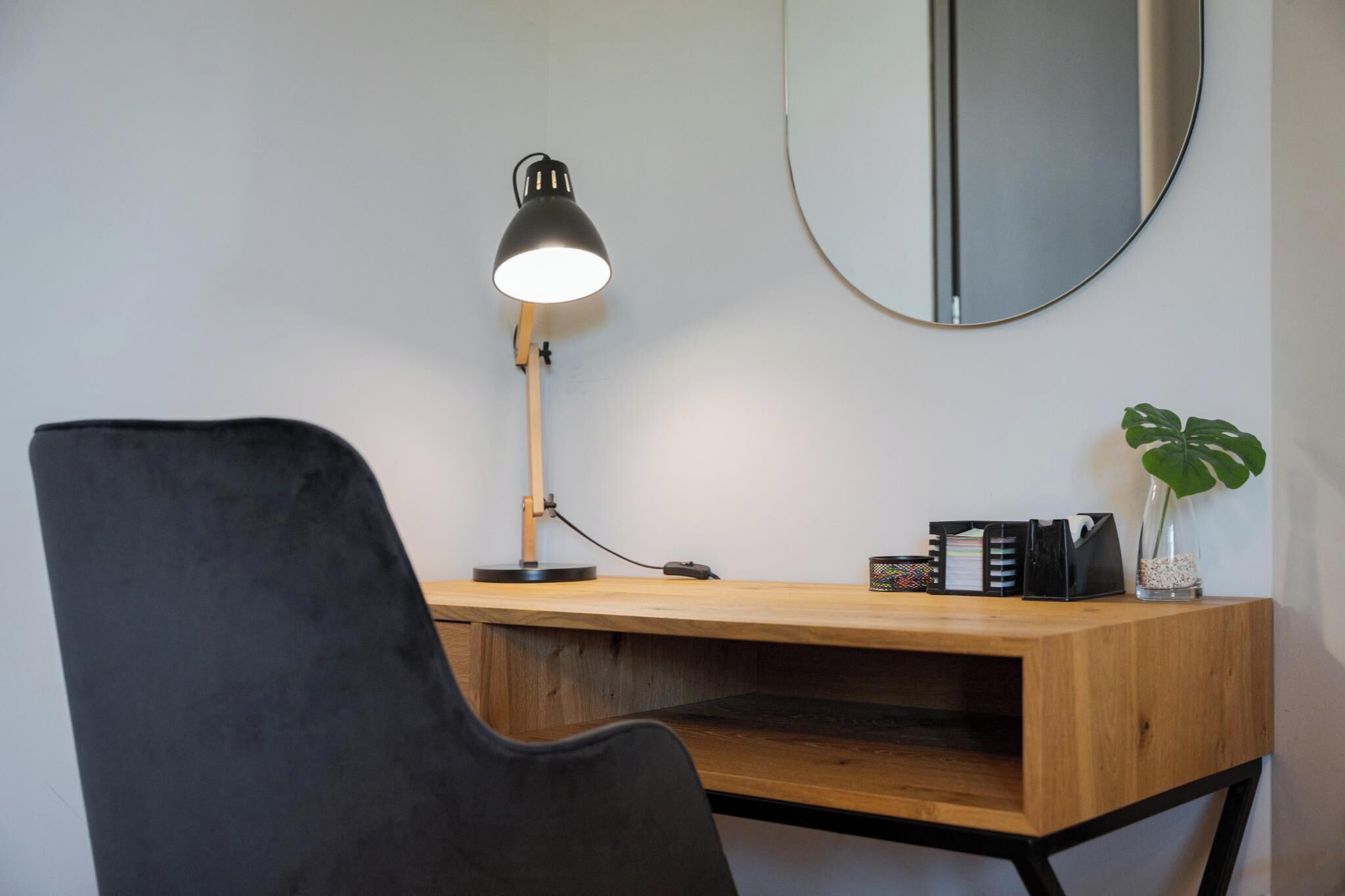 A wooden desk with a black chair, a desk lamp, a mirror, and a plant.