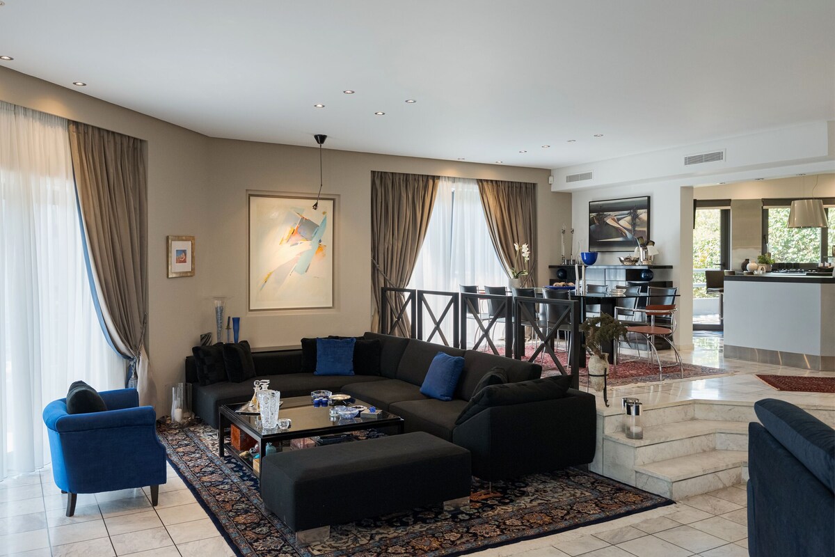 A modern living room with a black sofa, blue armchairs, and a coffee table. There is a dining area with a black table and chairs, and a kitchen visible in the background.