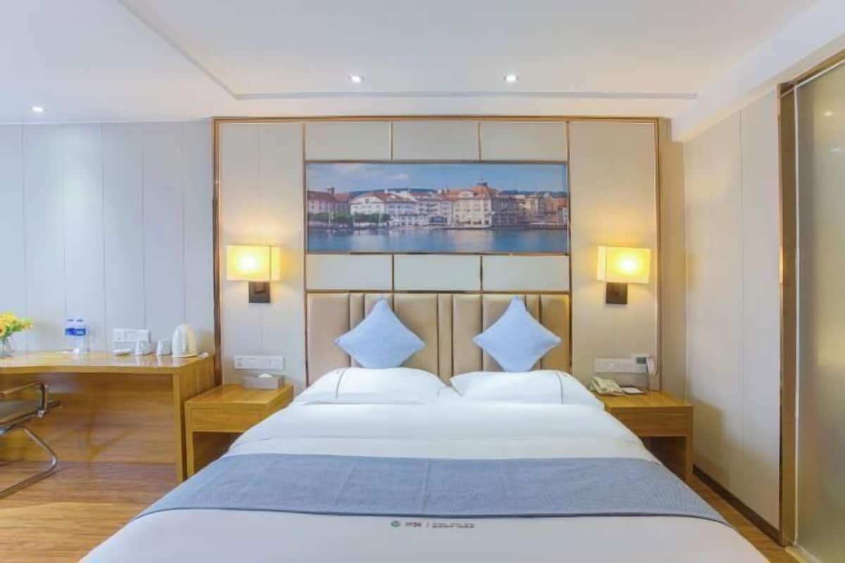 Une chambre d’hôtel avec un grand lit, deux tables de chevet, une tête de lit ornée d’un tableau représentant un paysage urbain et deux lampes fixées au mur.