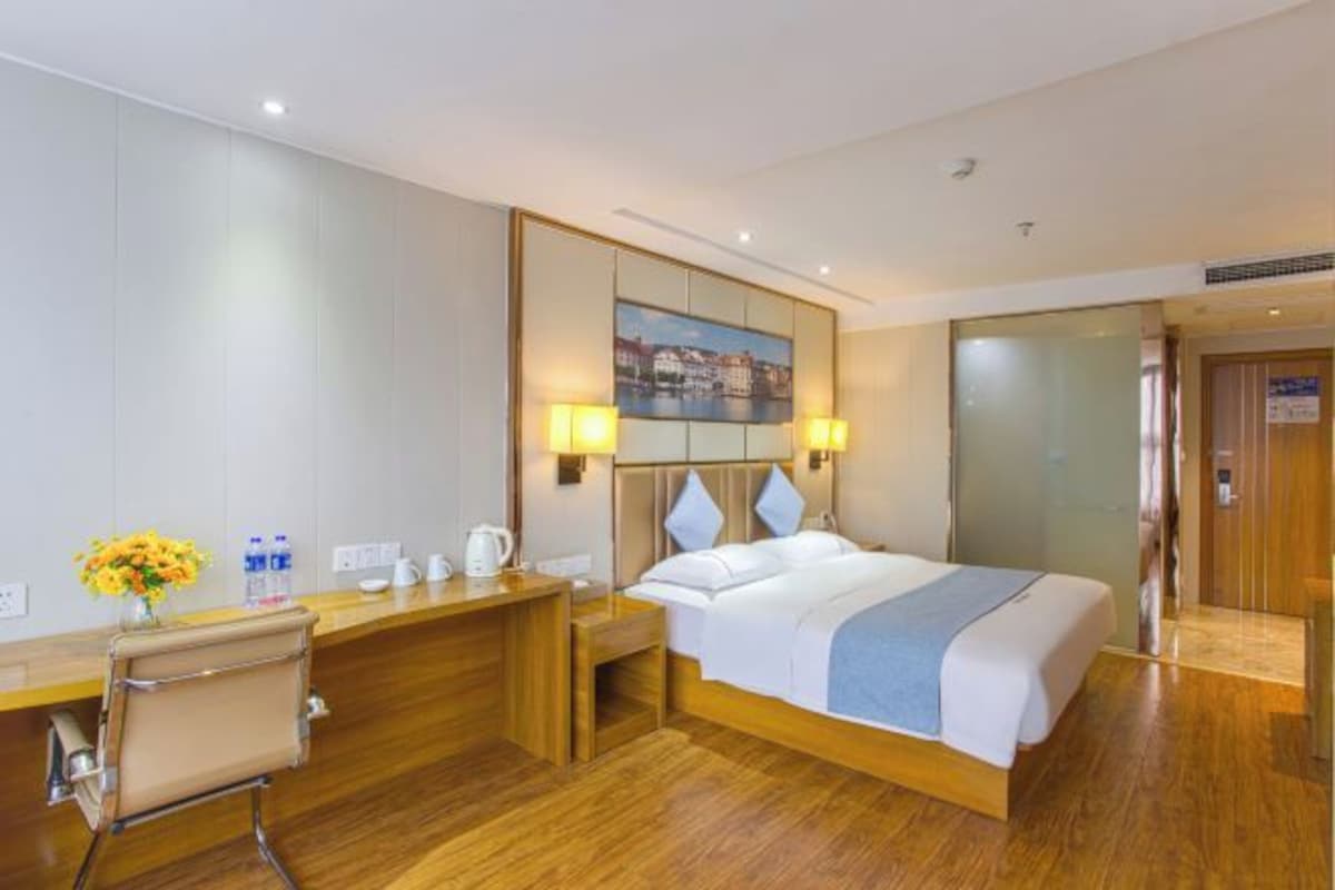 A hotel room with a bed, bedside table, lamp, and a painting on the wall.