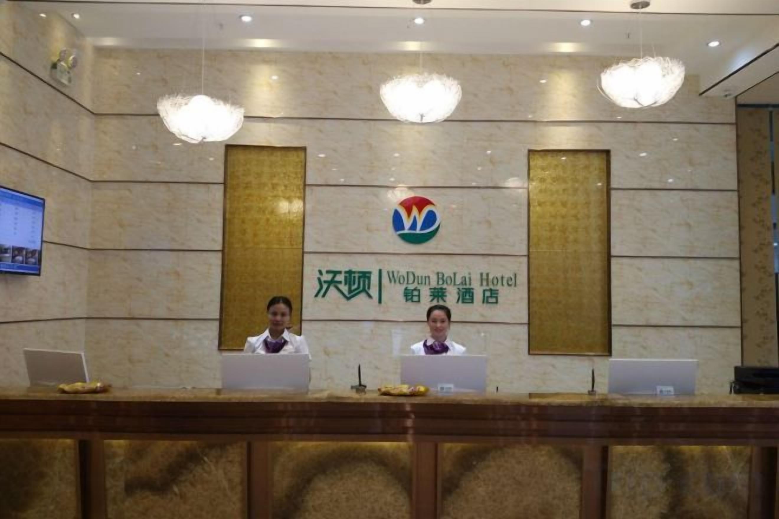 A reception desk at the WoDuBoLaI Hotel with two staff members behind it, a large logo above, and a television screen to the left.