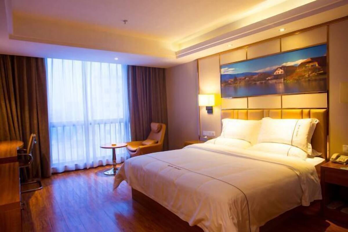 Une chambre d’hôtel avec un grand lit, une chaise, une petite table, un tableau au mur et une fenêtre avec des rideaux.