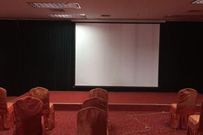 Sala de conferências com palco, tela de projetor e fileiras de cadeiras.