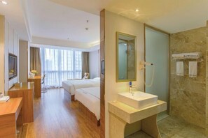 A hotel room with a wooden floor, a bed, a desk, a mirror, a sink, and a shower area.
