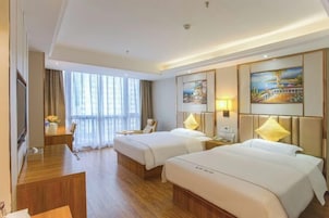 A hotel room with two beds, a desk, a chair, a TV, and large windows.