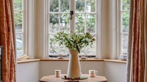 Une pièce avec une grande fenêtre, un vase blanc orné de fleurs posé sur une table en bois, et deux petites tasses.