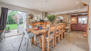 A spacious dining area with a wooden table and chairs, a kitchen in the background, and a view of the garden through an open door. - Impressive five bedroom country home in the Cotswolds - Barnsley Cottage (Cirencester)