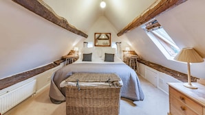 A bedroom with a sloped ceiling, exposed wooden beams, a bed with a headboard, a small table, a lamp, and a window. - Impressive five bedroom country home in the Cotswolds - Barnsley Cottage (Cirencester)