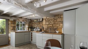 Une cuisine aux murs en pierre, avec un plan de travail en bois et un coin repas où une table est dressée pour deux personnes.