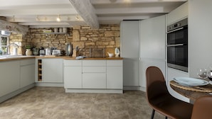 Une cuisine aux murs en pierre, avec des meubles blancs et un coin repas agrémenté d’une chaise marron.