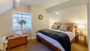 A bedroom with a bed, bedside table, lamp, and a window with flowers. - 18th century thatched Cotswold holiday cottage in Broadway - Orchard Cottage, Broadway (Broadway)
