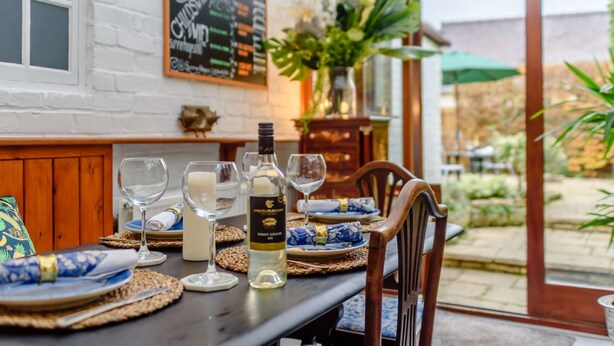 A dining table set with plates, glasses, and a bottle of wine, with a menu board and a vase of flowers in the background. - Family friendly period holiday cottage in the Cotswolds - Summer Hayes (Childswickham near Broadway)