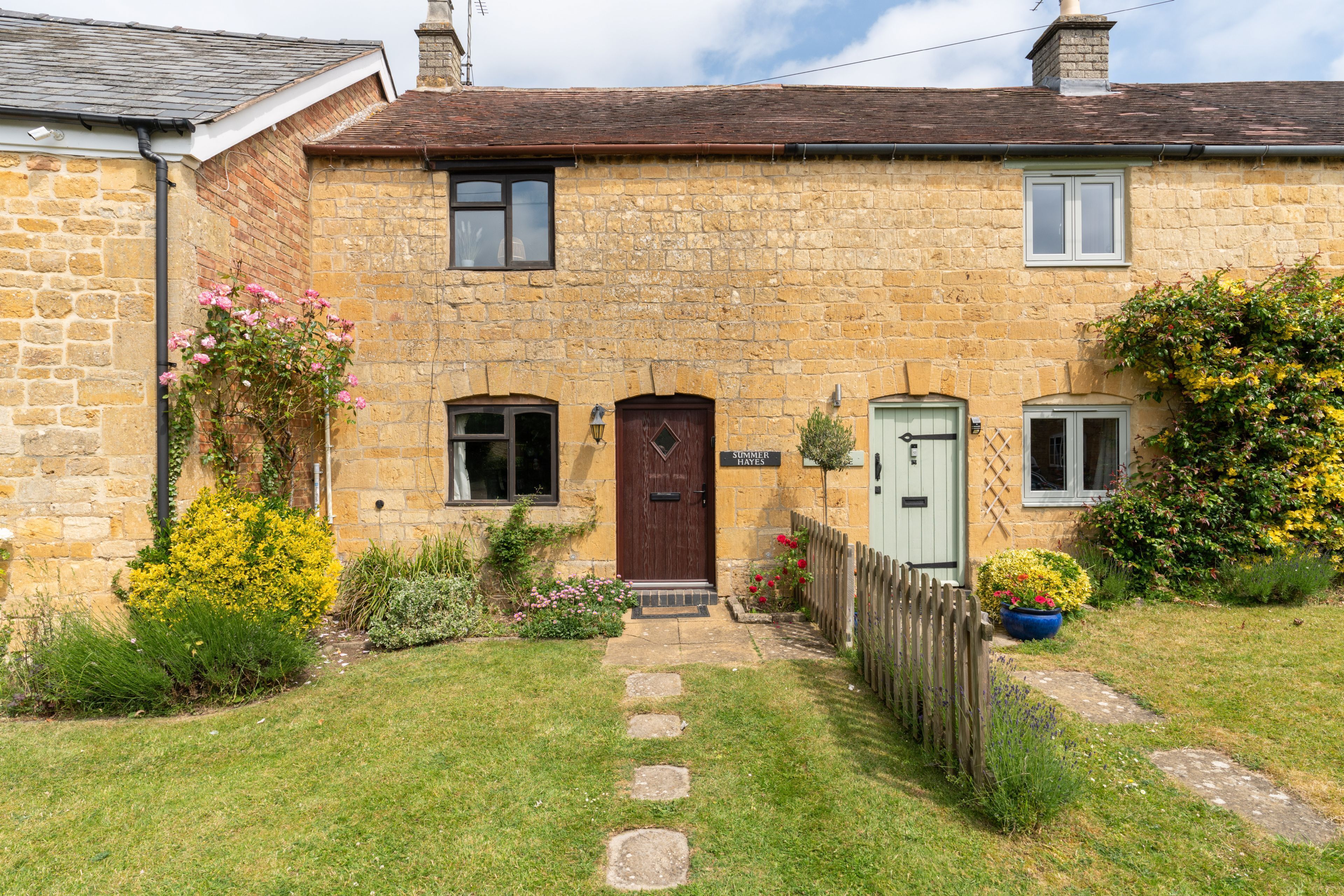A stone house with a wooden door, a small garden with flowers and a wooden fence.