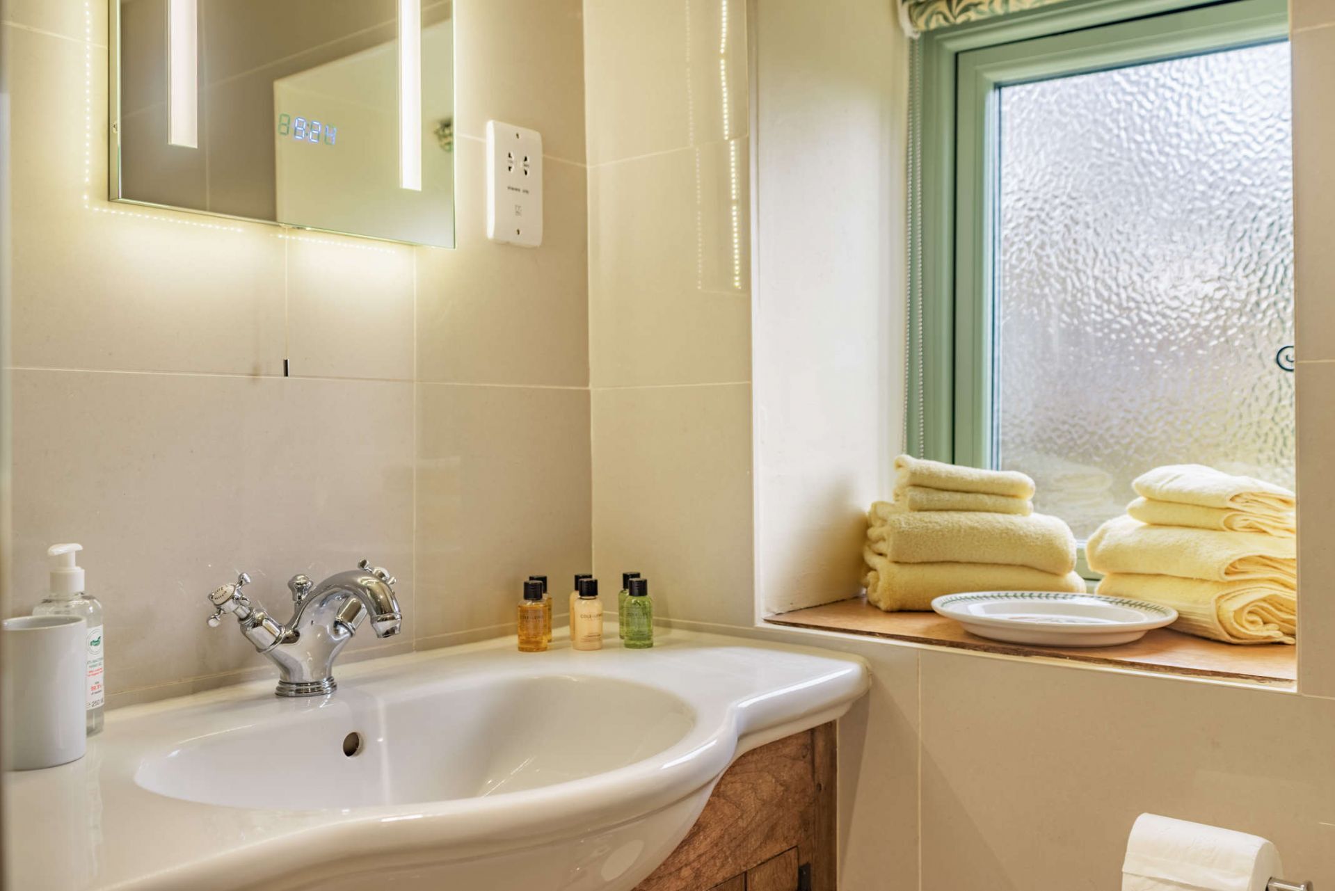 A bathroom with a sink, mirror, toiletries, and towels by a window.