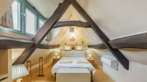 A cozy attic bedroom with exposed wooden beams, a bed with white linens, bedside lamps, and a small table with a lamp. - A delightful two bedroom Cotwold holiday cottage with garden - Cosy Corner (Moreton-in-Marsh)