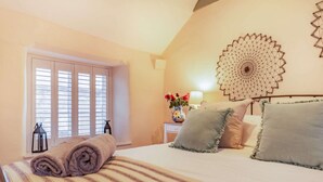 A bedroom with a bed, pillows, a bedside table with a lamp, a vase with flowers, and a wall hanging. - Dog friendly Cotswold cottage with a tranquil garden - Blenheim Cottage (Woodstock)