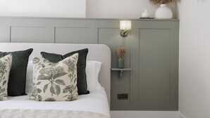 A bedroom with a bed, pillows, a nightstand, a wall-mounted lamp, and a vase with dried plants. - Elegant family friendly holiday cottage in the Cotswolds - The Smithy (Chipping Norton)