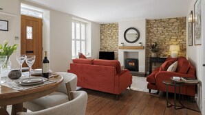A living room with a fireplace, red sofas, a round table, and a dining area with a wooden table and chairs. - Elegant family friendly holiday cottage in the Cotswolds - The Smithy (Chipping Norton)
