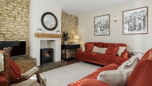 A cozy living room with a stone wall, a fireplace, a red sofa, and two framed black and white pictures. - Elegant family friendly holiday cottage in the Cotswolds - The Smithy (Chipping Norton)