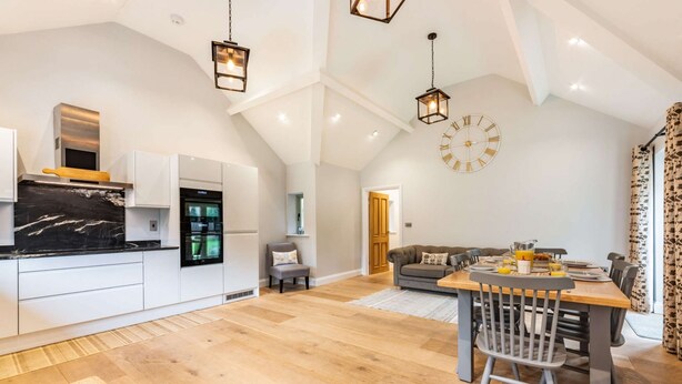 Un'ampia cucina con elettrodomestici moderni, una zona pranzo con tavolo e sedie in legno e un soggiorno con divano e un grande orologio a parete.
