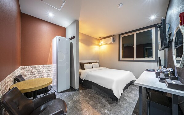 A compact hotel room with a bed, a small table, a chair, a kitchenette, and a window.
