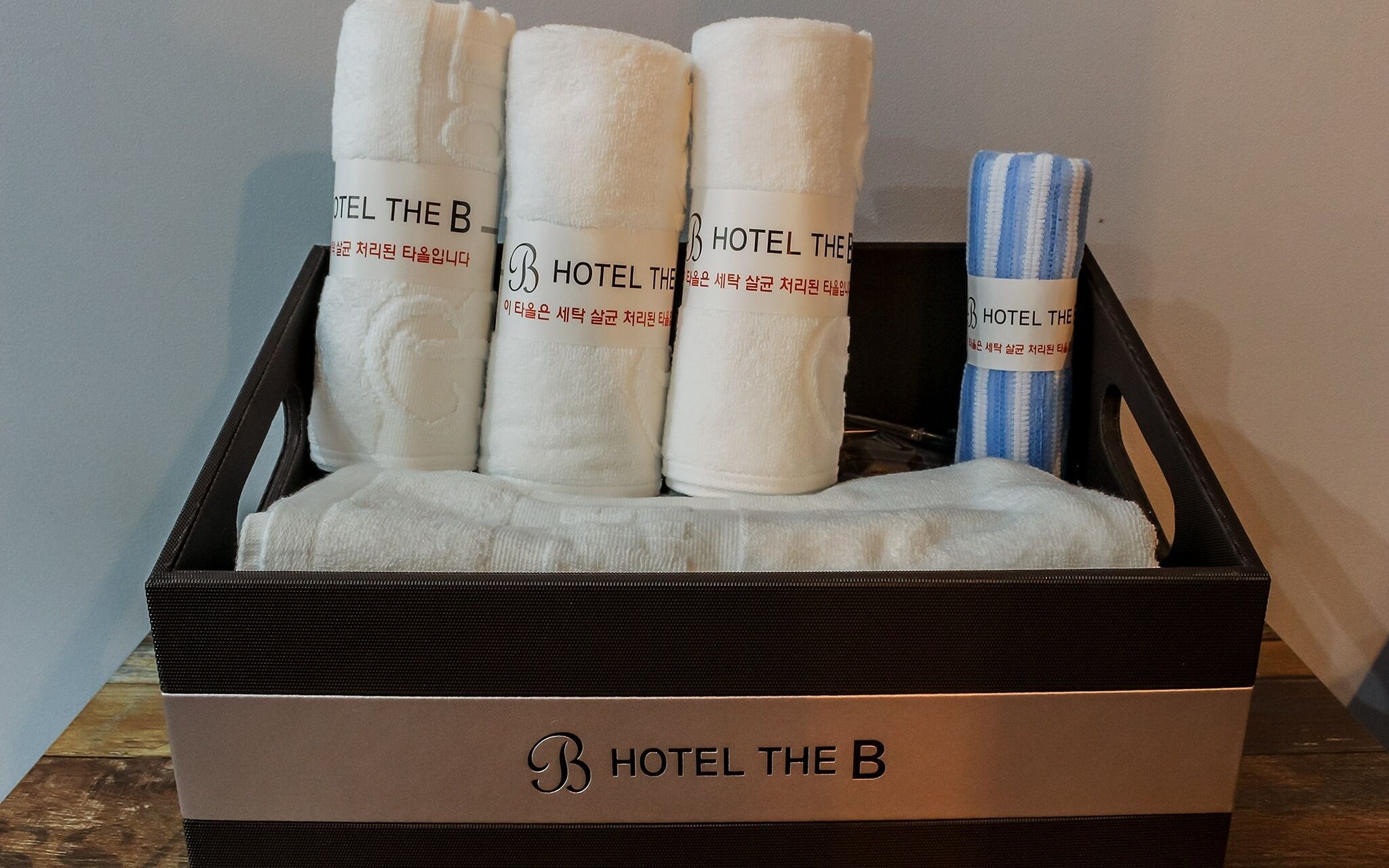 Une boîte noire ornée du logo « HOTEL THE B » contient quatre serviettes blanches avec du texte en coréen et une serviette bleue et blanche à rayures.