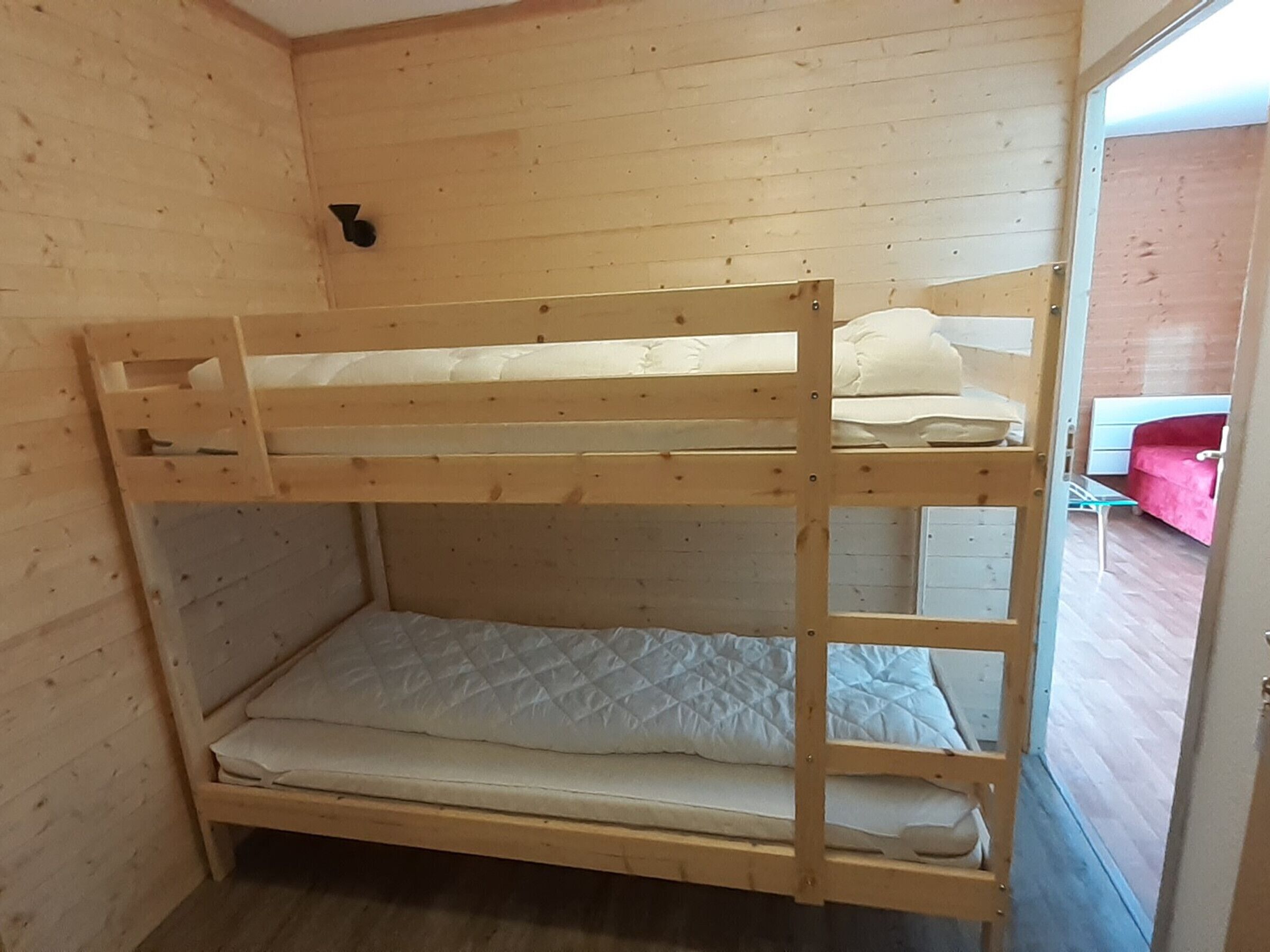 Un lit superposé avec une structure en bois et une literie blanche, dans une pièce aux murs en bois et dont on aperçoit la porte menant à une autre pièce.