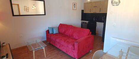 A compact living space with a red sofa, a small glass table, and a kitchenette in the background.