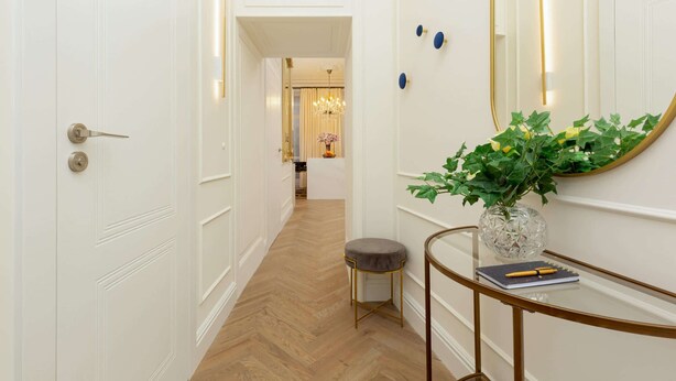 A hallway with a wooden floor, a white door, a small stool, a glass table with a vase and a notebook, and a mirror with a gold frame.